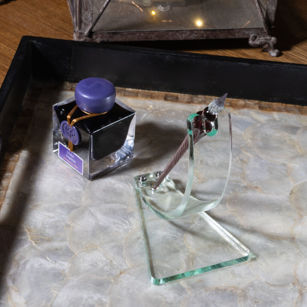 A green glass acrylic dip pen stand made with a curved strip bolted to a roughly trapezoidal base. A notch accommodates the shoulder of the pen at the top while a circular cut-out at the bottom keeps it from sliding. The elegran dip pen in the stand is violet with a clear tip. It sits next to a bottle of violet ink on a tray with a mother-of-pearl bottom and black sides.