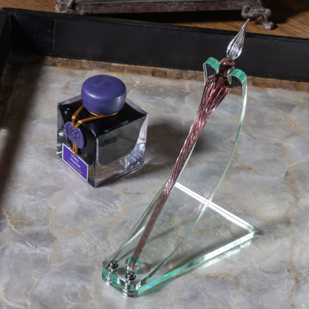 A green glass acrylic dip pen stand made with a curved strip bolted to a roughly trapezoidal base. A notch accommodates the shoulder of the pen at the top while a circular cut-out at the bottom keeps it from sliding. The elegran dip pen in the stand is violet with a clear tip. It sits next to a bottle of violet ink on a tray with a mother-of-pearl bottom and black sides.