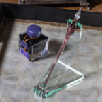 A green glass acrylic dip pen stand made with a curved strip bolted to a roughly trapezoidal base. A notch accommodates the shoulder of the pen at the top while a circular cut-out at the bottom keeps it from sliding. The elegran dip pen in the stand is violet with a clear tip. It sits next to a bottle of violet ink on a tray with a mother-of-pearl bottom and black sides.