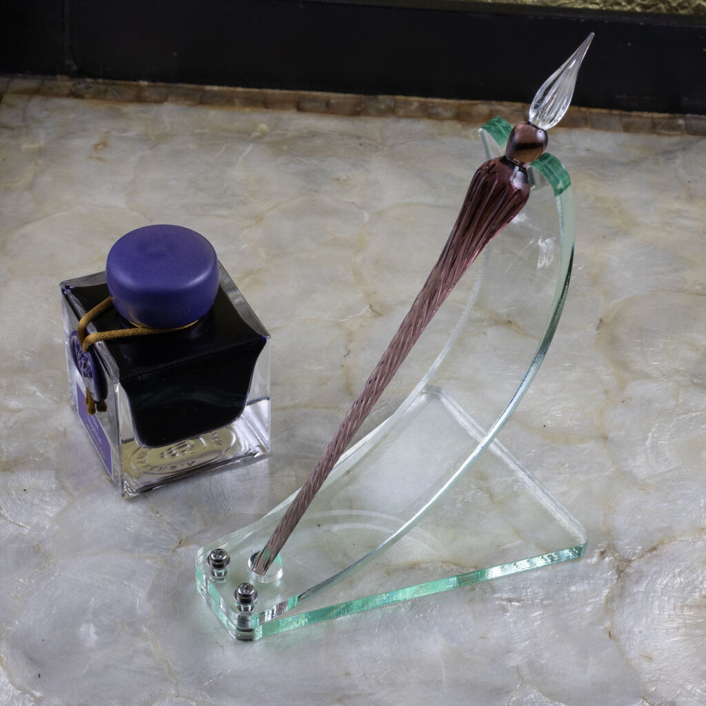 A green glass acrylic dip pen stand made with a curved strip bolted to a roughly trapezoidal base. A notch accommodates the shoulder of the pen at the top while a circular cut-out at the bottom keeps it from sliding. The elegran dip pen in the stand is violet with a clear tip. It sits next to a bottle of violet ink on a tray with a mother-of-pearl bottom and black sides.