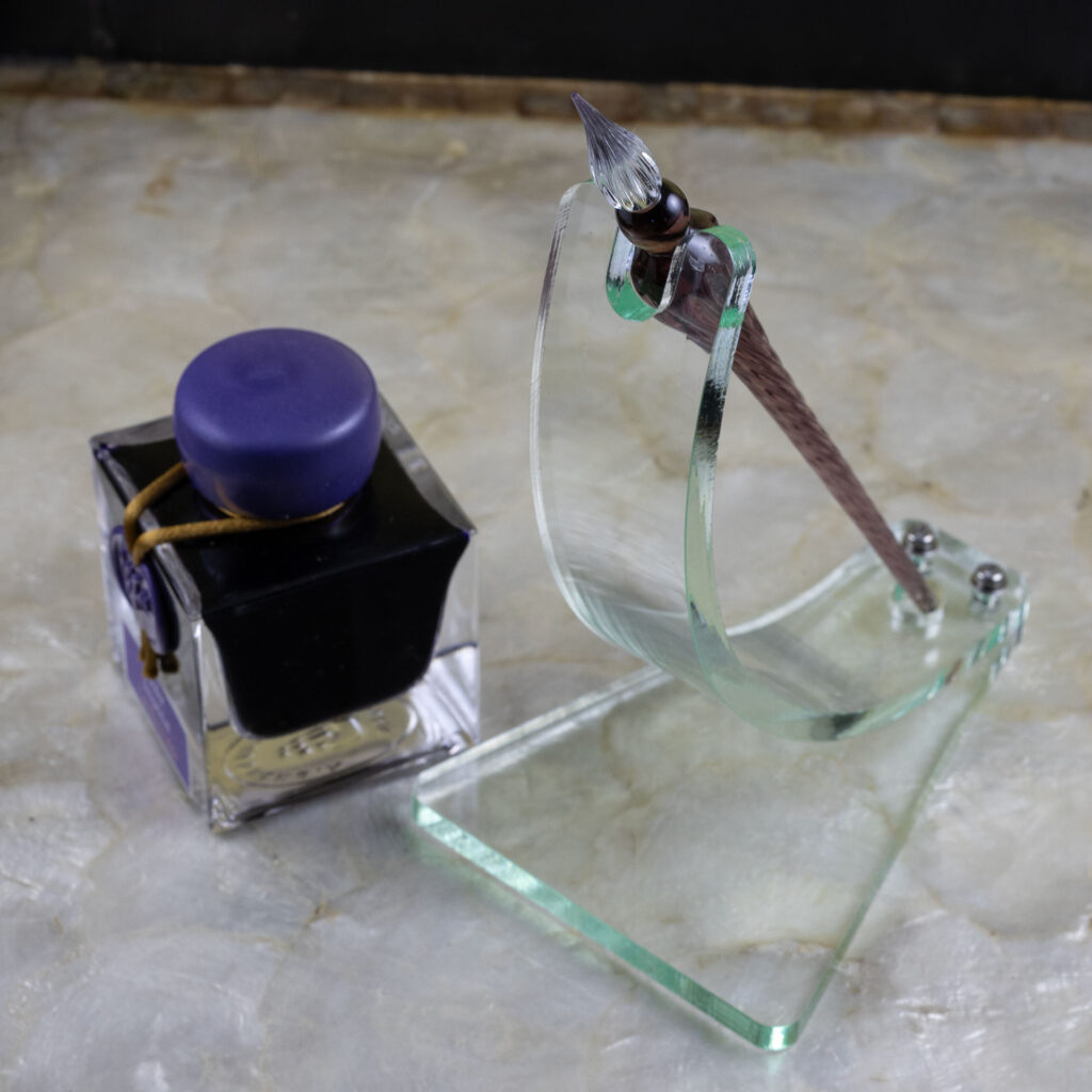 A green glass acrylic dip pen stand made with a curved strip bolted to a roughly trapezoidal base. A notch accommodates the shoulder of the pen at the top while a circular cut-out at the bottom keeps it from sliding. The elegran dip pen in the stand is violet with a clear tip. It sits next to a bottle of violet ink on a tray with a mother-of-pearl bottom and black sides.