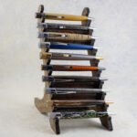 A pen rack made of walnut hardwood with two parallel angled vertical pieces connected by three flat cross members. The front-facing edges are serrated to hold 10 pens. The two front-facing cross members are inlaid with an off-center Celtic spiral made of padauk hardwood. The rack contains ten Retro 1951 pens (curvy, retro-future design shape with knurled twist-to-open tops, pen clips, in a variety of materials and finishes). Three-quarters left-side view.