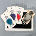 A rubber stamp of a line-art curvy potion bottle with a Celtic knotwork stopper lies face-up on a large black ink pad next to three cards with impressions from the stamp painted in with different inks. The first is a sparkly teal ink, the second is a dark violet-purple ink, and the last is bright red. Everything sits on the background of a grey self-healing cutting mat with a white grid.