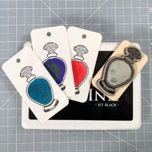 A rubber stamp of a line-art curvy potion bottle with a Celtic knotwork stopper lies face-up on a large black ink pad next to three cards with impressions from the stamp painted in with different inks. The first is a sparkly teal ink, the second is a dark violet-purple ink, and the last is bright red. Everything sits on the background of a grey self-healing cutting mat with a white grid.