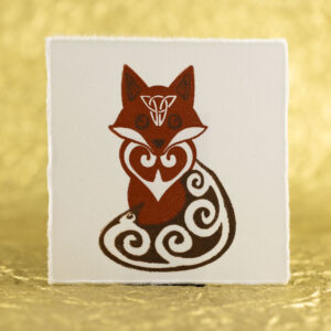 A fox with Celtic styling (spirals in the tail, ears, eyes, and chest with an inverted triangular knot on the forehead) printed in two shades of brown on a square of white paper with slightly ragged edges against a crinkled, defocused gold background.