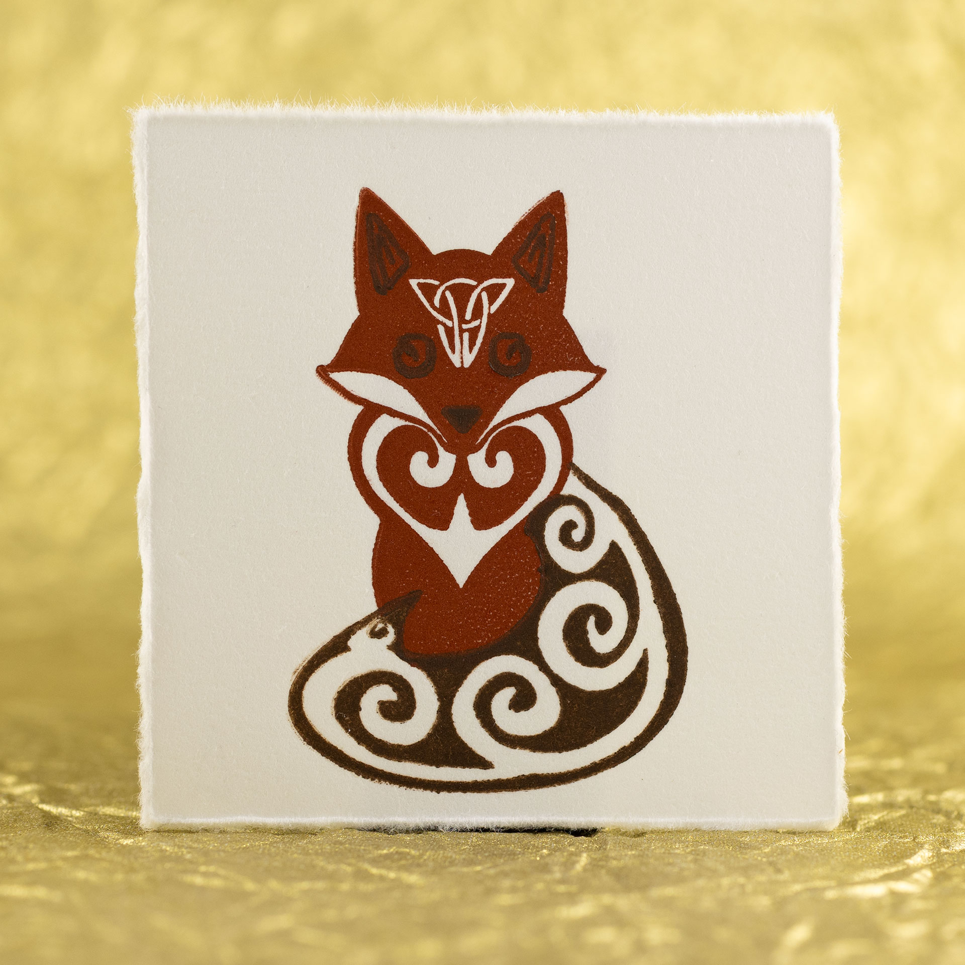 A fox with Celtic styling (spirals in the tail, ears, eyes, and chest with an inverted triangular knot on the forehead) printed in two shades of brown on a square of white paper with slightly ragged edges against a crinkled, defocused gold background.