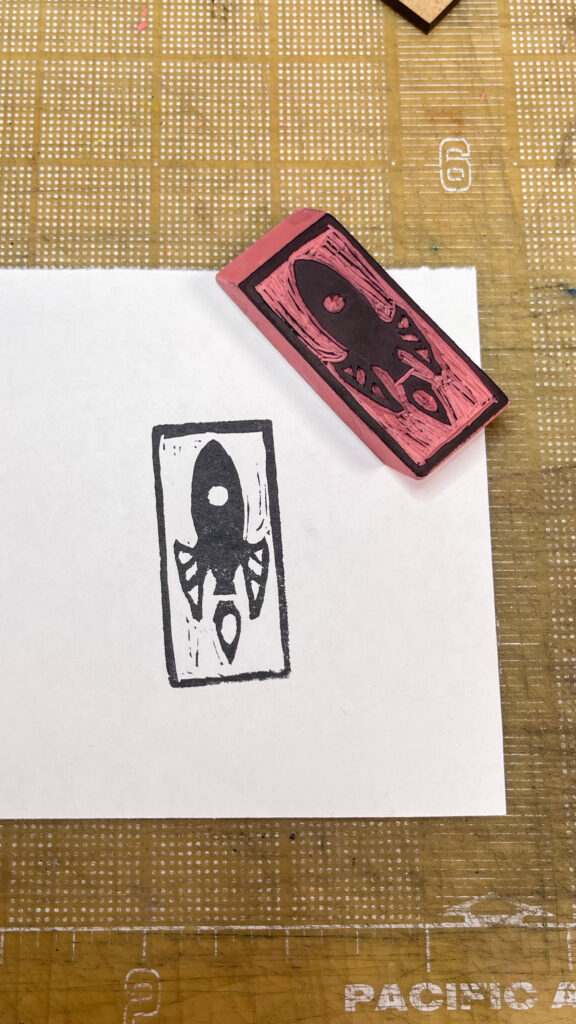 A rectangular pink rubber eraser with a retro-style rocket ship carved onto one face, the design highlighted in black laying face-up next to an imprint from the rocket eraser stamp on a piece of paper.