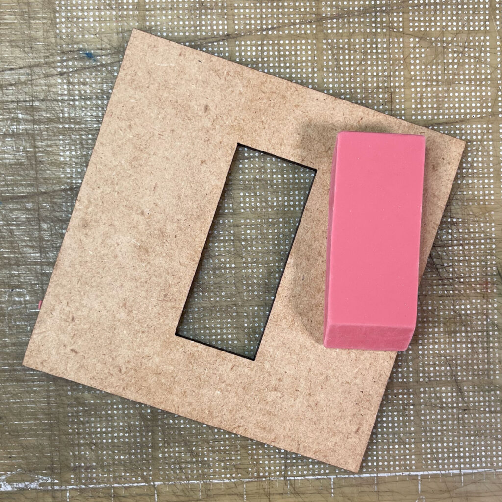 A square of 3mm thick MDF board with a rectangular hole about the size of the pink eraser sitting next to it.