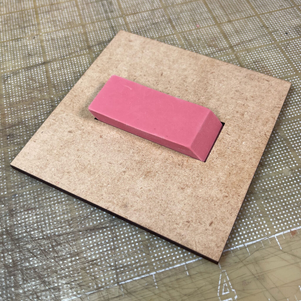 A square of 3mm thick MDF board with a rectangular hole with a pink rubber eraser pressed into it.