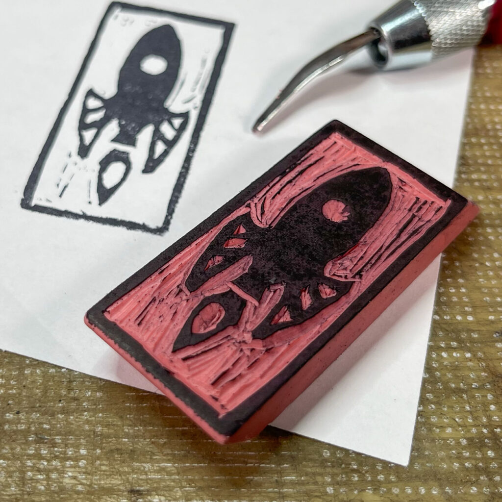 A rectangular pink rubber eraser with a retro-style rocket ship carved onto one face laying on a piece of paper that has been imprinted with the stamp. The tip of a lino carving tool is also visible.