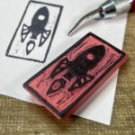 A rectangular pink rubber eraser with a retro-style rocket ship carved onto one face laying on a piece of paper that has been imprinted with the stamp. The tip of a lino carving tool is also visible.