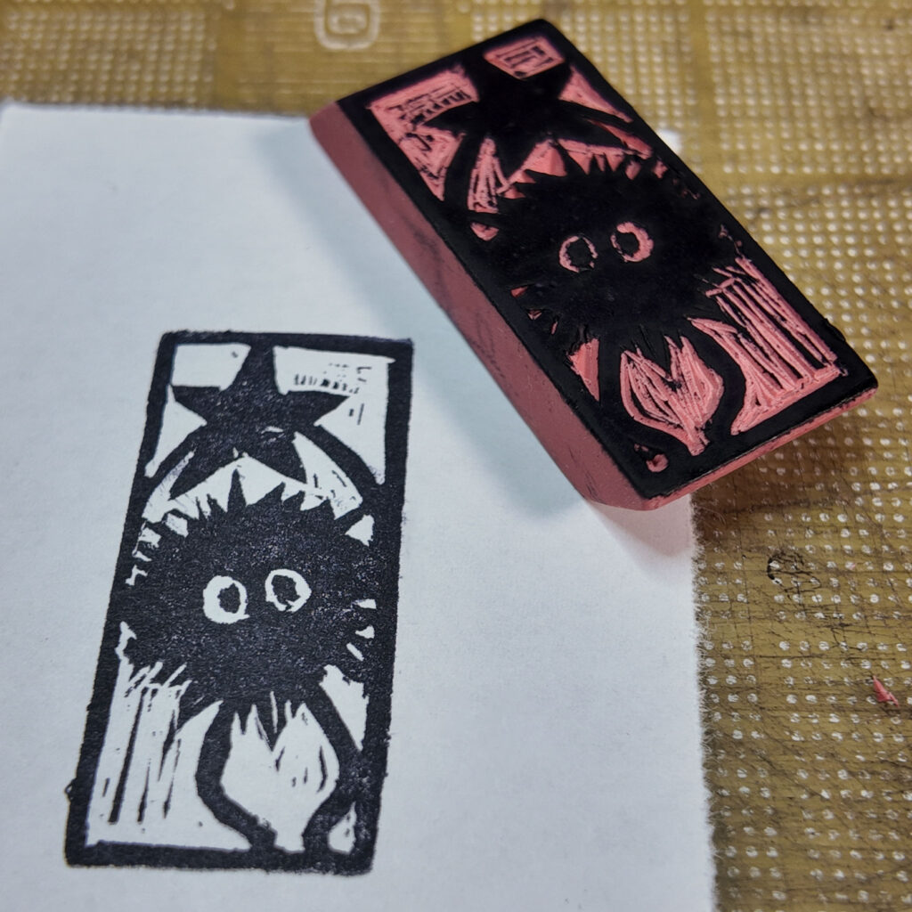 A rectangular pink rubber eraser with a soot sprite ship carved onto one face, the design highlighted in black laying face-up next to an imprint from the stamp on a piece of paper. The sprite is a cartoonish character, a fuzzy black ball with two simple eyes, and legs and arms that are simple curved lines. It is holding up a five-pointed star over its head.