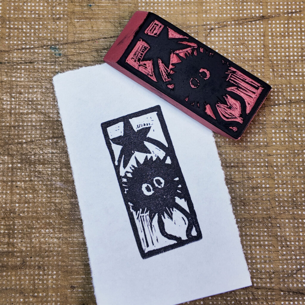 A rectangular pink rubber eraser with a soot sprite ship carved onto one face, the design highlighted in black laying face-up next to an imprint from the stamp on a piece of paper. The sprite is a cartoonish character, a fuzzy black ball with two simple eyes, and legs and arms that are simple curved lines. It is holding up a five-pointed star over its head.