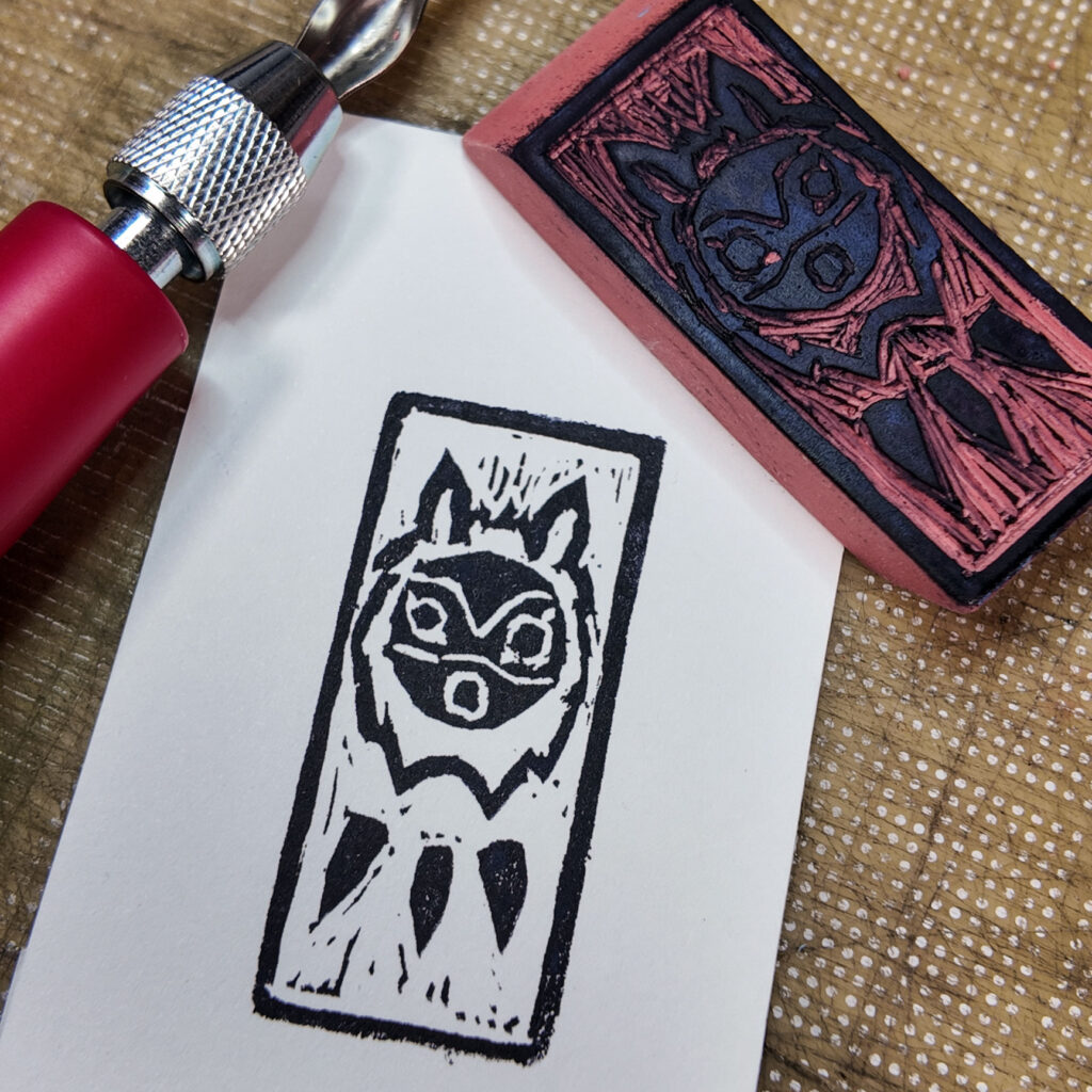 A stylized wolf mask from the animated movie Princess Mononoke with a rectangular border has been stamped onto a rectangle of paper. The mask is a circle with circles for eyes and mouth. A couple of curved lines across the mask hint at eyebrows and nose. The circular face is surrounded by a shaggy mane and topped by two animal ears. Below the mask are three pointy objects reminiscent of horns on a necklace. The carved eraser stamp that made the impression is face up on one corner of the print. A red-handled lino cutting knife and little bits of cut rubber are also visible.