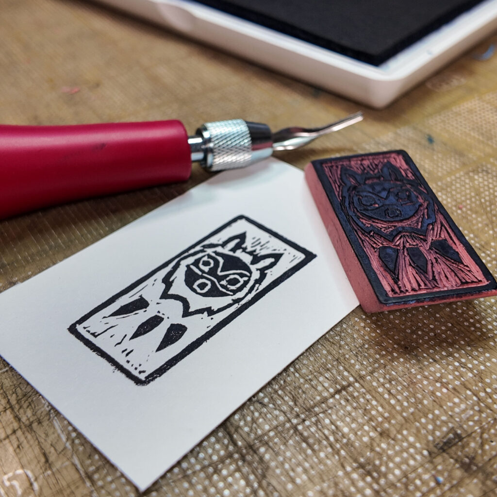 A stylized wolf mask from the animated movie Princess Mononoke with a rectangular border has been stamped onto a rectangle of paper. The mask is a circle with circles for eyes and mouth. A couple of curved lines across the mask hint at eyebrows and nose. The circular face is surrounded by a shaggy mane and topped by two animal ears. Below the mask are three pointy objects reminiscent of horns on a necklace. The carved eraser stamp that made the impression is face up on one corner of the print. A red-handled lino cutting knife and the corner of a stamp pad are also visible.