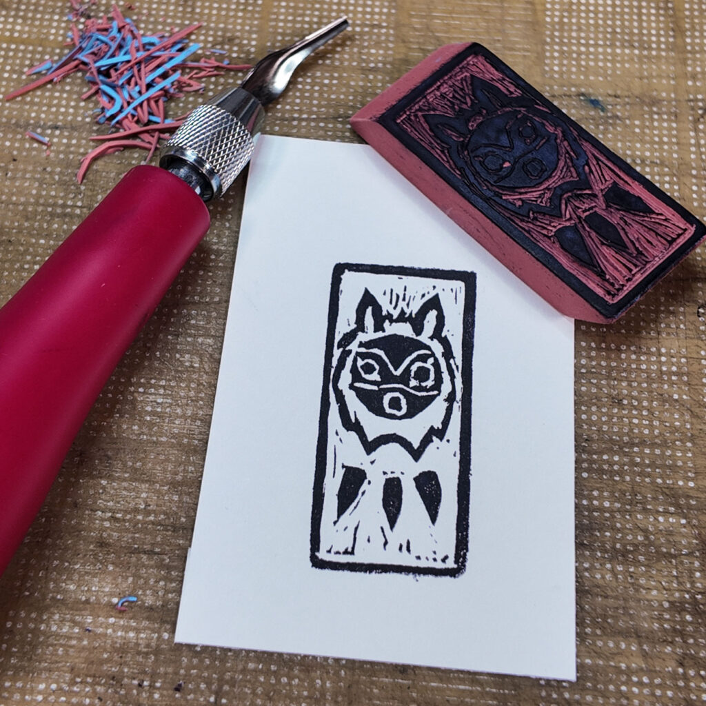A stylized wolf mask from the animated movie Princess Mononoke with a rectangular border has been stamped onto a rectangle of paper. The mask is a circle with circles for eyes and mouth. A couple of curved lines across the mask hint at eyebrows and nose. The circular face is surrounded by a shaggy mane and topped by two animal ears. Below the mask are three pointy objects reminiscent of horns on a necklace. The carved eraser stamp that made the impression is face up on one corner of the print. A red-handled lino cutting knife and little bits of cut rubber are also visible.