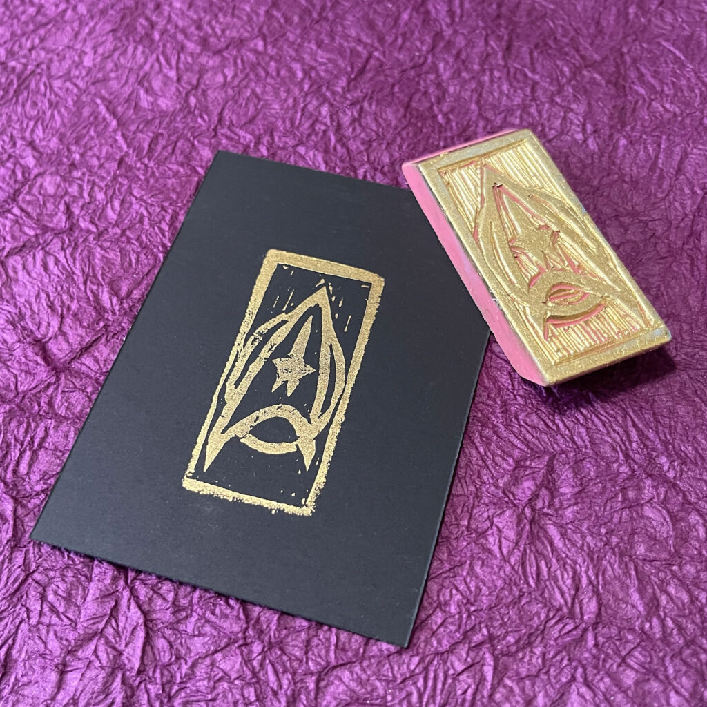 A Star Trek command insignia stamped in gold on black paper. The hand-carved eraser stamp that made the impression lays at an angle across the upper right corner of the paper.