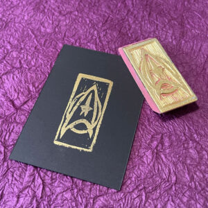 A Star Trek command insignia stamped in gold on black paper. The hand-carved eraser stamp that made the impression lays at an angle across the upper right corner of the paper.