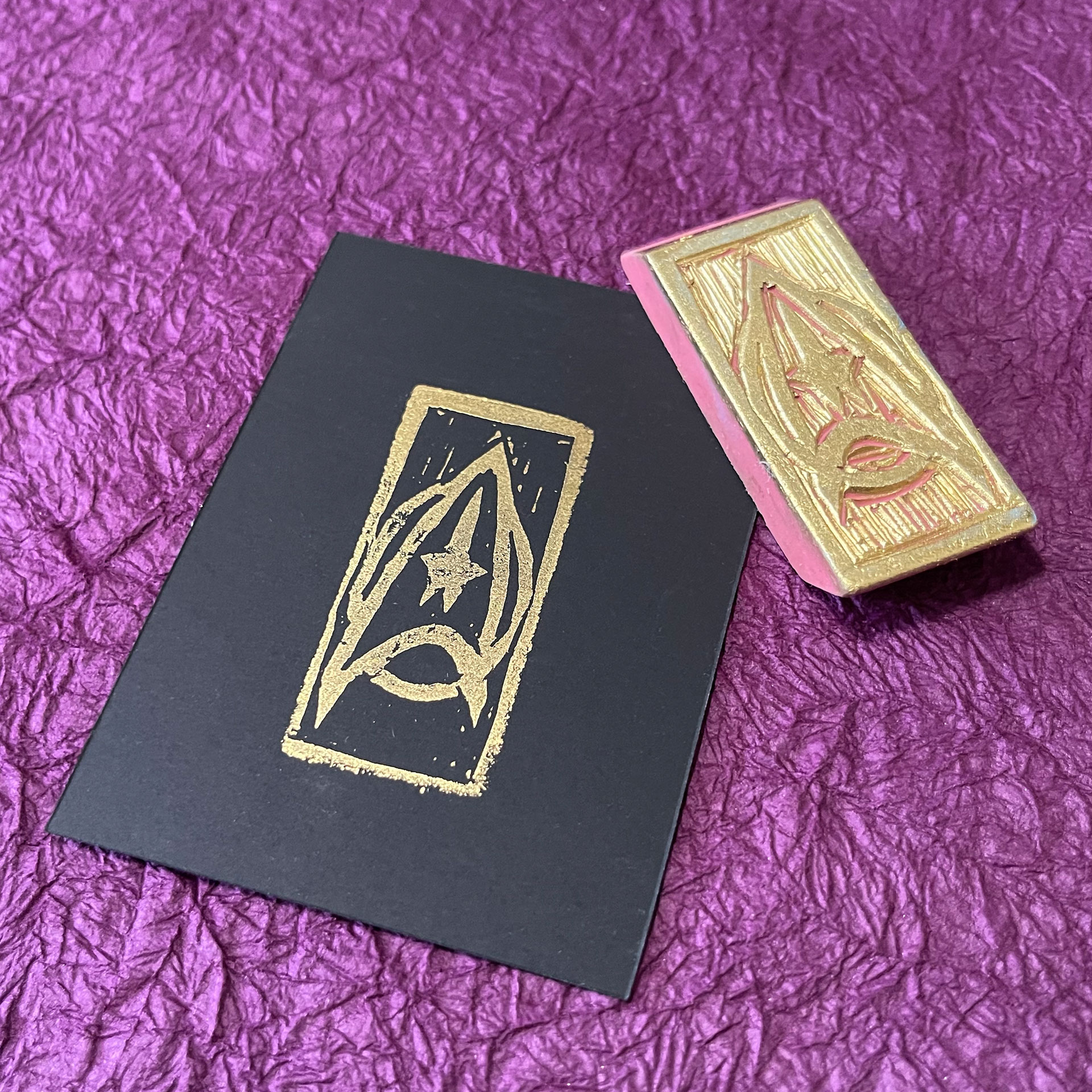 A Star Trek command insignia stamped in gold on black paper. The hand-carved eraser stamp that made the impression lays at an angle across the upper right corner of the paper.
