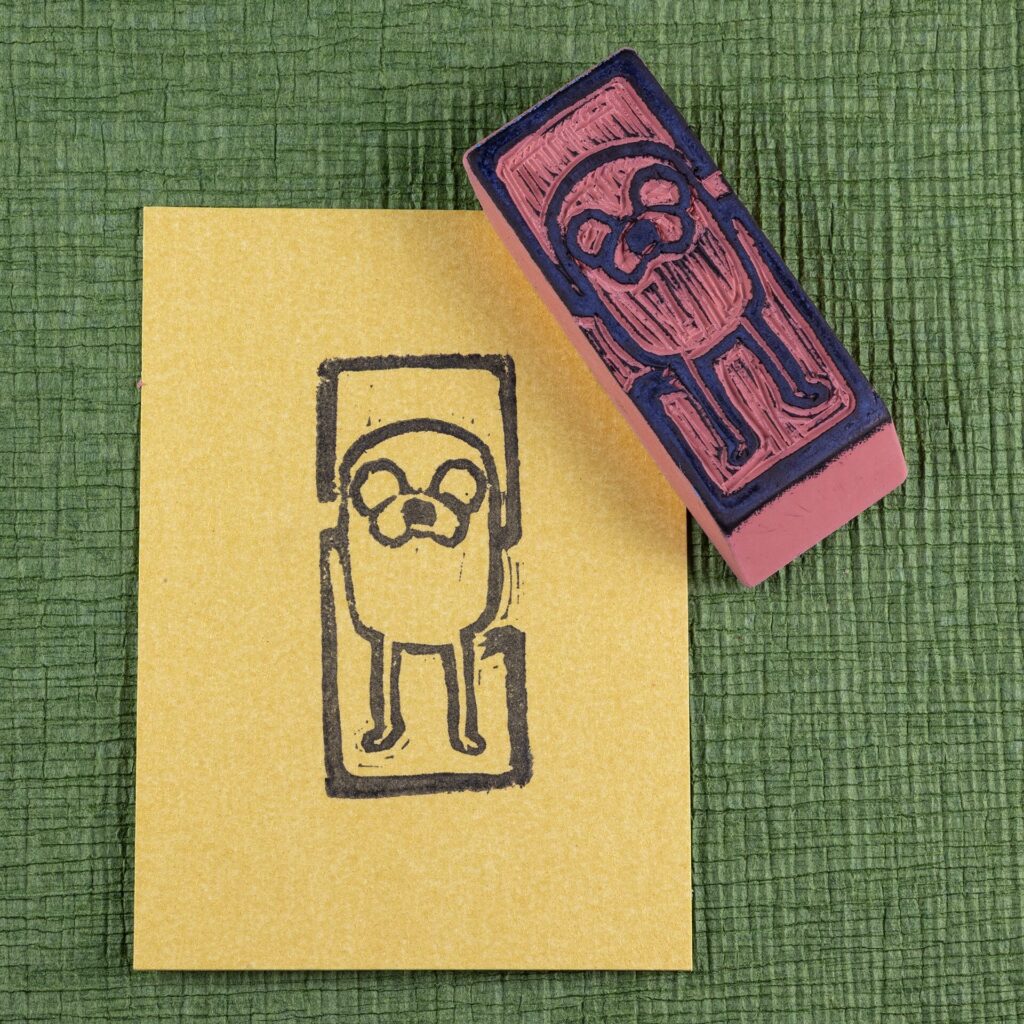 An eraser stamp carved with an image of Jake the Dog from the Adventure Time animated series is positioned face-up across the upper right corner of a rectangle of pearlescent yellow/gold stock with an impression from the stamp in black. Both sit on a green crepe surface (green chirimen paper). Top-down view.