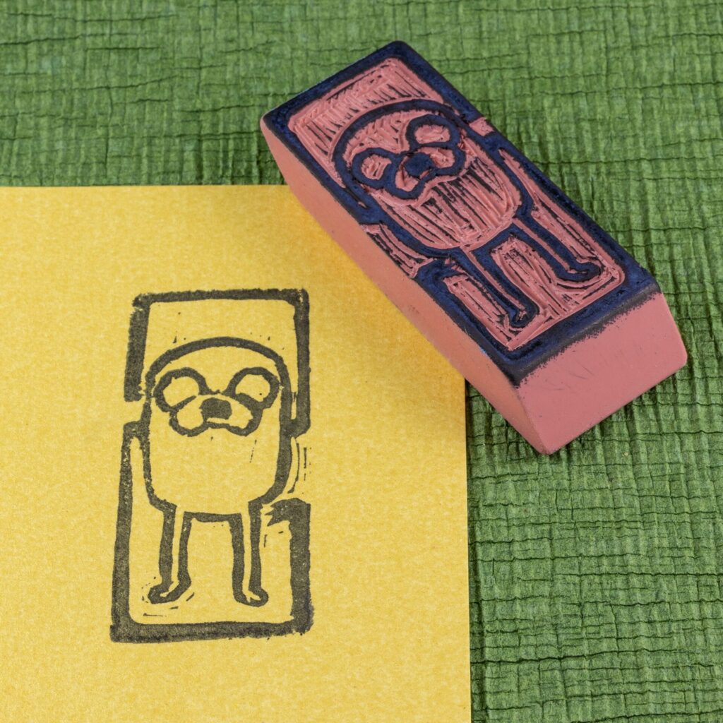 An eraser stamp carved with an image of Jake the Dog from the Adventure Time animated series is positioned face-up across the upper right corner of a rectangle of pearlescent yellow/gold stock with an impression from the stamp in black. Both sit on a green crepe surface (green chirimen paper). View from the top, angled from the lower left.