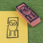 An eraser stamp carved with an image of Jake the Dog from the Adventure Time animated series is positioned face-up across the upper right corner of a rectangle of pearlescent yellow/gold stock with an impression from the stamp in black. Both sit on a green crepe surface (green chirimen paper). View from the top, angled from the lower left.