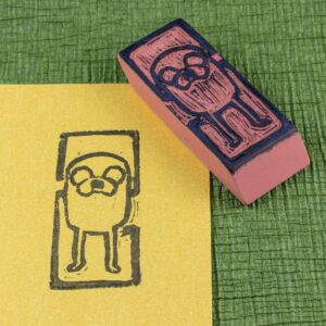 An eraser stamp carved with an image of Jake the Dog from the Adventure Time animated series is positioned face-up across the upper right corner of a rectangle of pearlescent yellow/gold stock with an impression from the stamp in black. Both sit on a green crepe surface (green chirimen paper). View from the top, angled from the lower left.