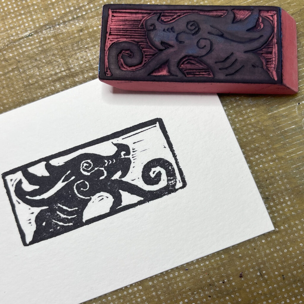 An eraser carved with a stylized dragon head featuring spirals for tongue, eye, nostril, and cheek sits on the upper right corner of a rectangle of white paper with an impression of the stamp in black. They are sitting on a well-weathered cutting mat.