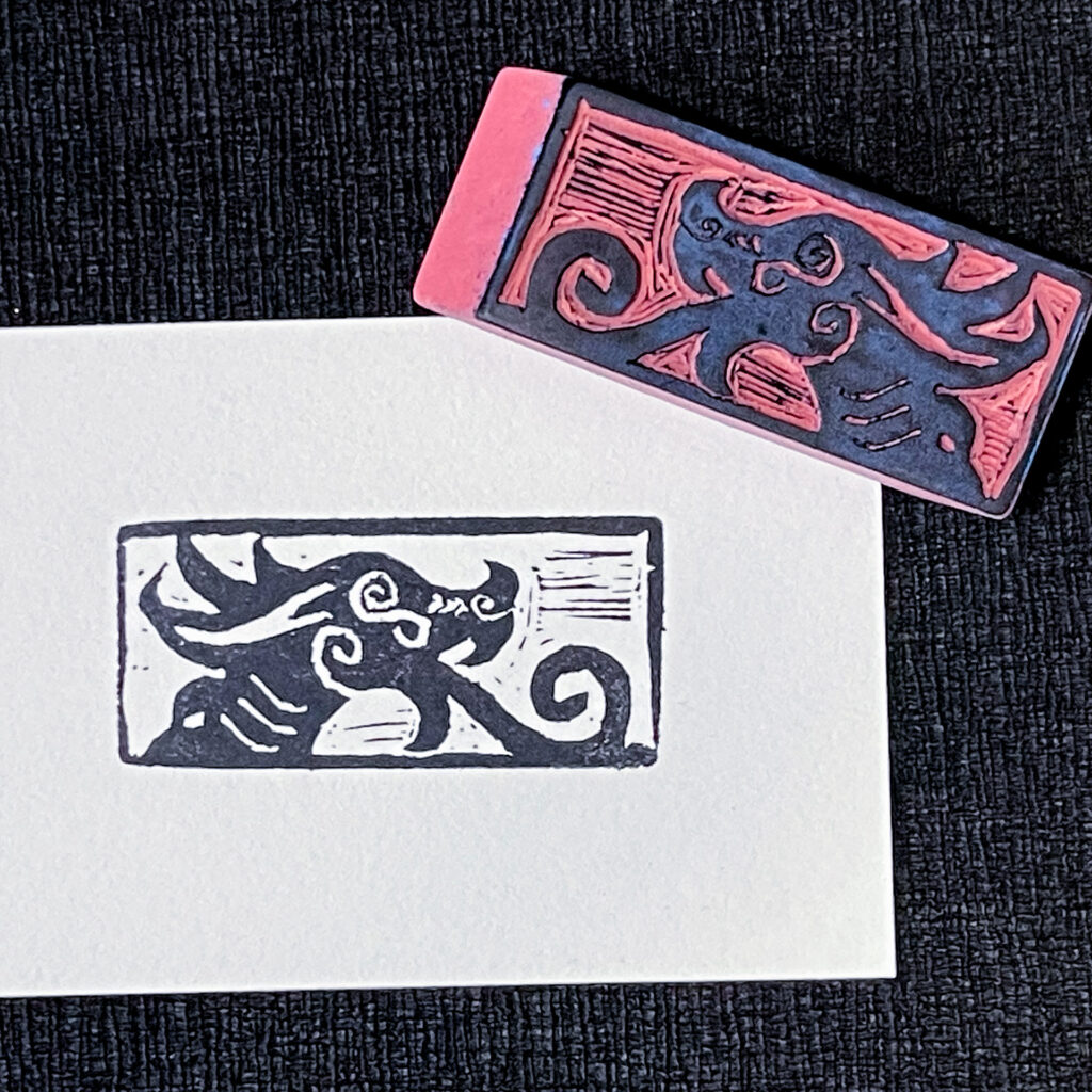 An eraser carved with a stylized dragon head featuring spirals for tongue, eye, nostril, and cheek sits on the upper right corner of a rectangle of white paper with an impression of the stamp in black. Both sit on a background of textured black chirimen paper.