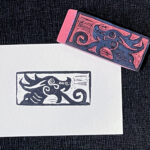 An eraser carved with a stylized dragon head featuring spirals for tongue, eye, nostril, and cheek sits on the upper right corner of a rectangle of white paper with an impression of the stamp in black. Both sit on a background of textured black chirimen paper.