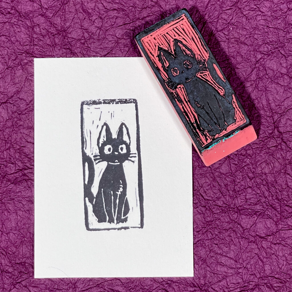 A white rectangle of paper imprinted with a cartoon-style black cat facing the viewer, its tail curling off to the left. The hand-carved eraser that made the imprint sits face up on the upper right corner of the paper. Both are on a background of crinkled purple paper.