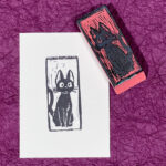 A white rectangle of paper imprinted with a cartoon-style black cat facing the viewer, its tail curling off to the left. The hand-carved eraser that made the imprint sits face up on the upper right corner of the paper. Both are on a background of crinkled purple paper.