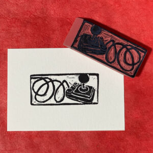 A rectangular piece of white paper in landscape format is imprinted in black with and angled view of an old-style home computer joystick. The cord from the joystick coils irregularly away from the joystick, then turns into a rectangular border around the print. The carved eraser that made the print it face-up on the upper right corner of the print. Both are sitting on a surface of bright red paper.
