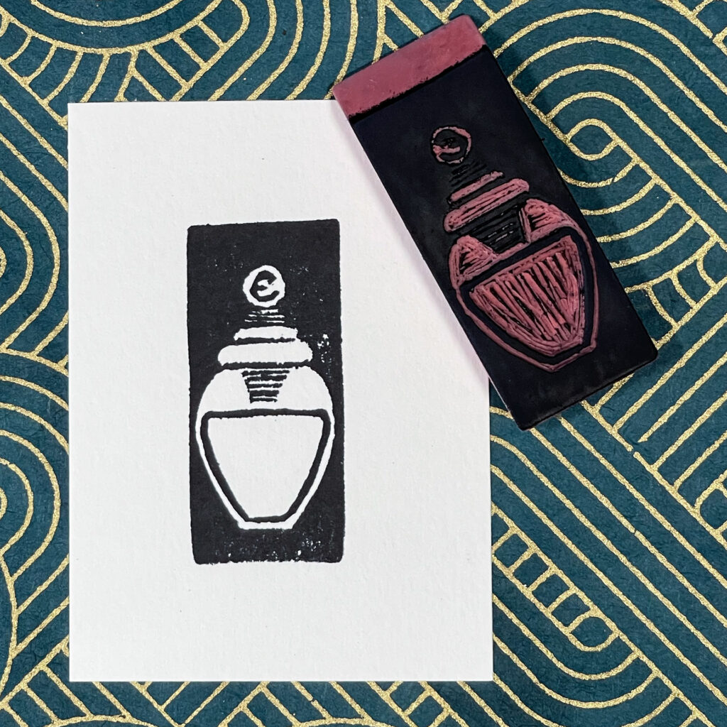 A rectangular piece of paper is imprinted in black with an image of a small jar of ink. The carved eraser stamp that made the impression lays face-up on the upper right corner of the print. The background is blue-green decorative paper with gold metallic lines. Straight-on view.