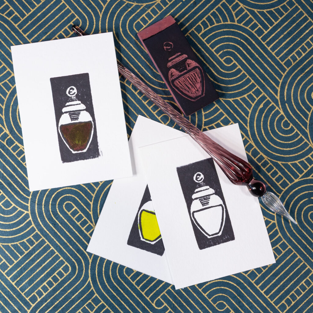Several rectangular pieces of paper imprinted in black with images of a small jar of ink. They overlap some. The body of one jar has been painted in with a dark ink with a green sheen. Another has been filled with a yellow-green. The third is un-colored. A purple glass did pen lays across the prints. The carved eraser that made the prints is face-up in the upper right. The background is blue-green decorative paper with gold metallic lines.