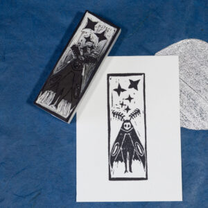 A rectangular piece of white paper imprinted with an anthropomorphized, cartoonish moth character with five four-pointed stars in various sizes above its head. The wings look like a cloak around a very simple body. The face is white with two black spots for eyes. There are feathered antennae atop the head. The blocky white carved eraser that made the impression is laying face-up on the upper left corner of the print. Both are on a background of deep blue paper with a single metallic silver leaf inclusion.