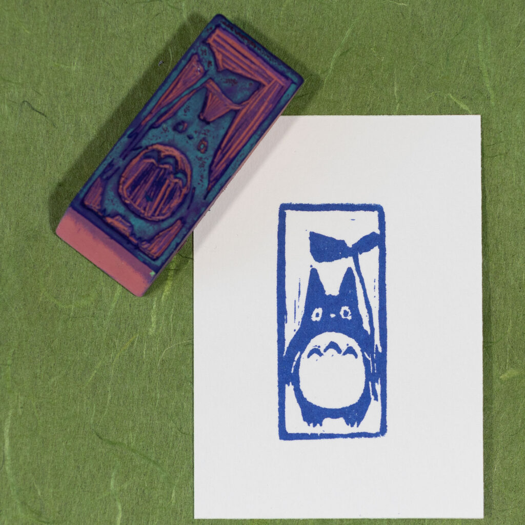 A rectangle of white paper imprinted in blue with the older child totoro character from the animated movie My Neighbor Totoro. It has a stout, round body with a big round white patch on its stomach. There are three chevrons on its chest. It has stubby legs and arms, long pointed ears, and big oval eyes. It is holding a leaf with a long stem as an umbrella. The carved eraser that made the print is laying at an angle on the upper left corner. The eraser and print are both sitting on a sheet of handmade grass green paper with long fibrous inclusions in different shades of green.