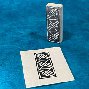 A blocky white eraser has been carved with a Celtic key pattern, a maze-like pattern consisting of angled lines and triangles. The raised surface of the design is stained black. The stamp stands on one small edge on a surface of blue handmade paper. In front of the stamp is a rectangular piece of white paper that has been imprinted with the design in black.