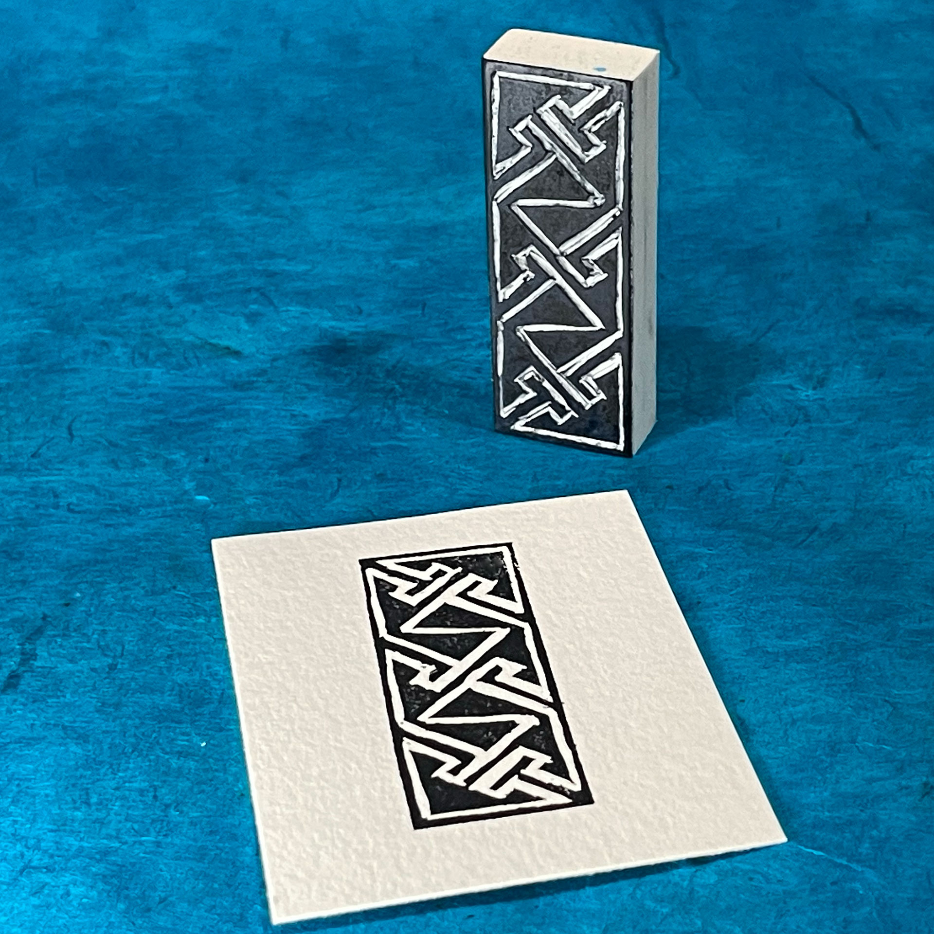 A blocky white eraser has been carved with a Celtic key pattern, a maze-like pattern consisting of angled lines and triangles. The raised surface of the design is stained black. The stamp stands on one small edge on a surface of blue handmade paper. In front of the stamp is a rectangular piece of white paper that has been imprinted with the design in black.