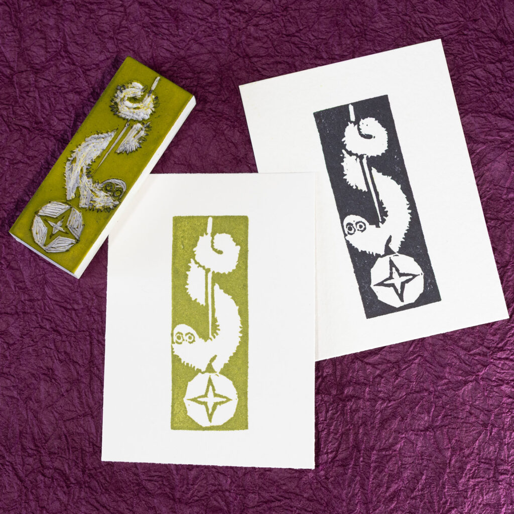 A couple of white rectangles of paper are imprinted with an image of long fuzzy cartoon creature wrapped around a cord suspending a ball with a four-pointed star. The image is reversed out of the ink making up the print. There is one print in an avocado green and another in black. The carved white eraser (stained with green ink) rests face-up on the corner of the green print. The background is crinkled purple paper (momigami).