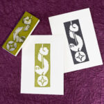 A couple of white rectangles of paper are imprinted with an image of long fuzzy cartoon creature wrapped around a cord suspending a ball with a four-pointed star. The image is reversed out of the ink making up the print. There is one print in an avocado green and another in black. The carved white eraser (stained with green ink) rests face-up on the corner of the green print. The background is crinkled purple paper (momigami).
