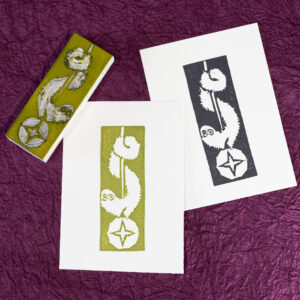 A couple of white rectangles of paper are imprinted with an image of long fuzzy cartoon creature wrapped around a cord suspending a ball with a four-pointed star. The image is reversed out of the ink making up the print. There is one print in an avocado green and another in black. The carved white eraser (stained with green ink) rests face-up on the corner of the green print. The background is crinkled purple paper (momigami).