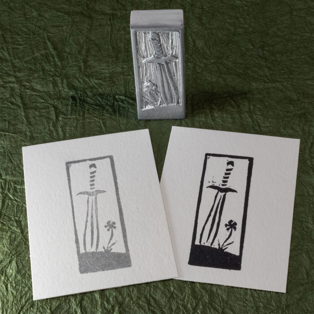 A stamp hand-carved from a blocky white eraser stands on one of the narrow sides on a background of crinkled dark green paper (momigami) behind two impressions made from it, one in black ink, one in silver ink. The design is a leaf-blade sword with the tip stuck into the ground next to a single flower.