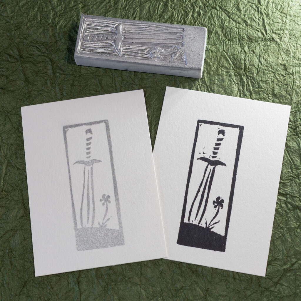A stamp hand-carved from a blocky white eraser lays face-up on a background of crinkled dark green paper (momigami) behind two impressions made from it, one in black ink, one in silver ink. The design is a leaf-blade sword with the tip stuck into the ground next to a single flower.