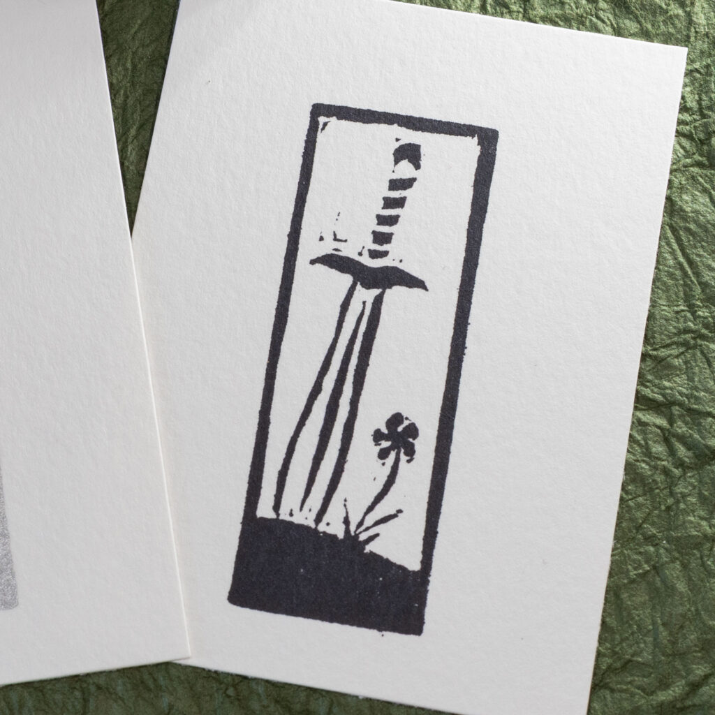 Eraser stamp imprint of a leaf-blade sword with the tip stuck into the ground next to a single flowef in black ink on white paper.