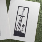 Eraser stamp imprint of a leaf-blade sword with the tip stuck into the ground next to a single flowef in black ink on white paper.