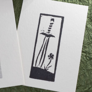 Eraser stamp imprint of a leaf-blade sword with the tip stuck into the ground next to a single flowef in black ink on white paper.