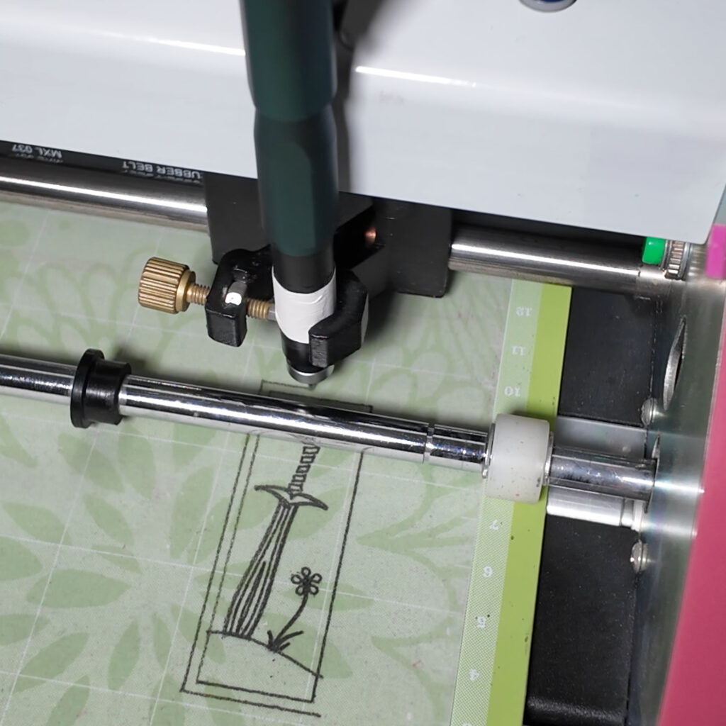 Close-up of a Bosskut Gazelle plotting cutter. There is a mechanical pencil in the tool holder, and a rectangle with a sword and flower are being drawn on a piece of tracing paper on the cutting mat.
