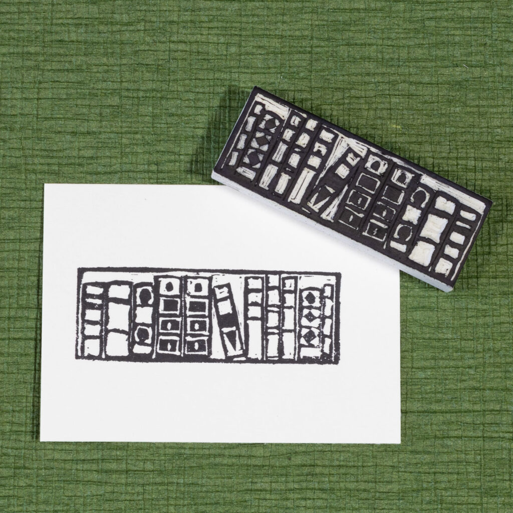 A white rectangle of paper imprinted with the image of a bookshelf with eleven books of different sizes. One book in the middle is tilted to the left within a larger gap. The hand-carved white blocky eraser that made the impression is laying face-up across the upper right corner. Both are on a surface of dark green crepe paper (chirimen).