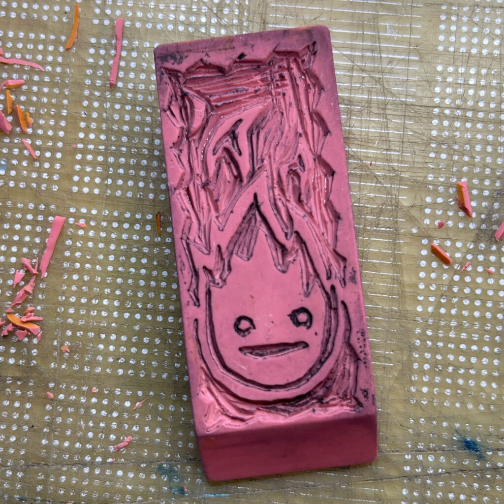 Cleaned off hand-carved Calcifer pink eraser stamp. Calcifer is a stylized flame with two simple eyes and a mouth.